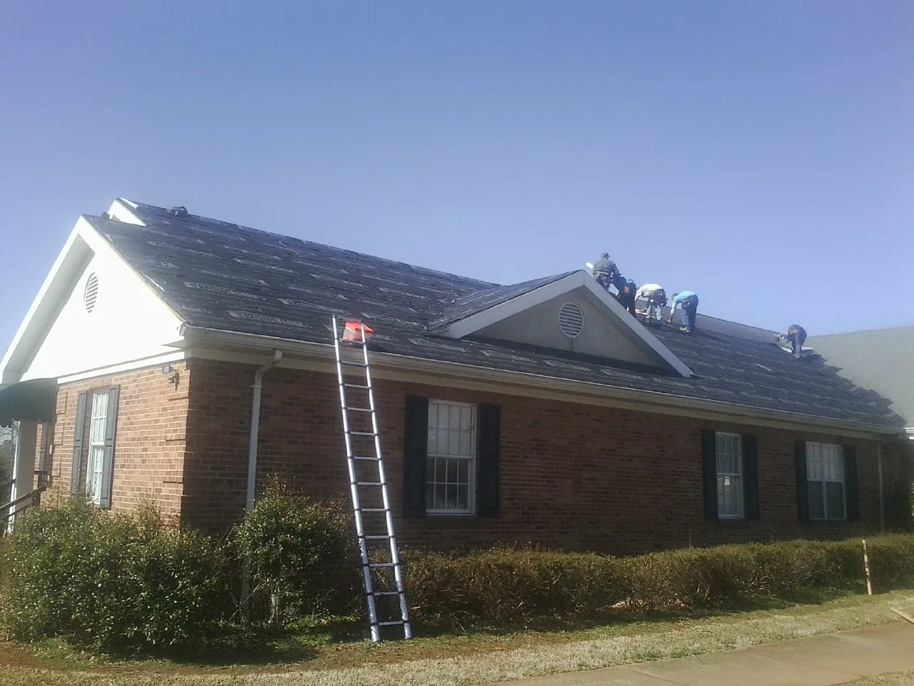 Crew tearing shingles off a brick ranch roof with a ladder set — Metal Roof Repair work by our crew in Newry