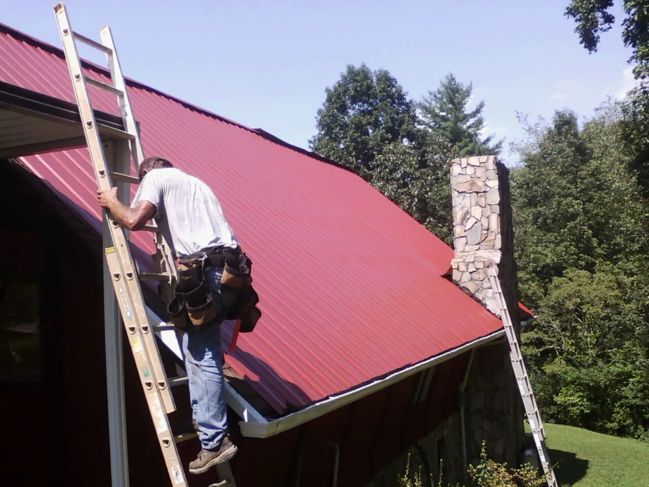 Roofer on a ladder beside a red metal roof and stone chimney — Soffit & Fascia Repair by Roofing Brothers in Newry